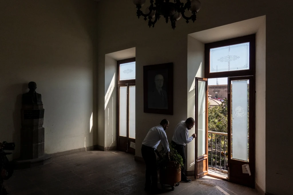 Dwie osoby w białych koszulach stoją przy otwartym oknie w jasnym pomieszczeniu, w tle widoczne popiersie oraz duży portret na ścianie.