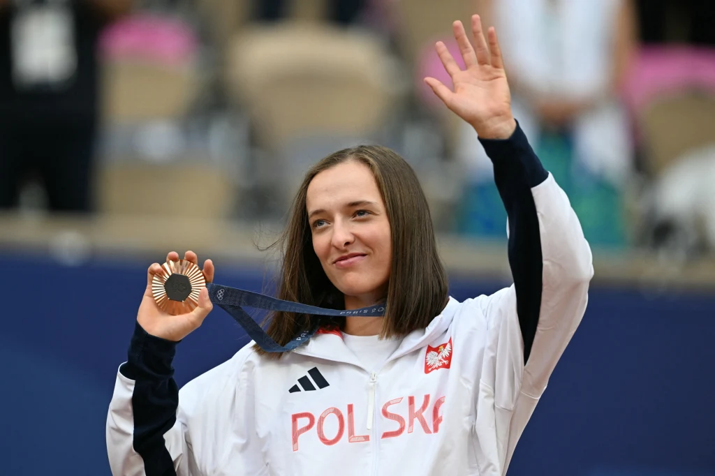 Iga Świątek z brązowym medalem igrzysk olimpijskich Sportowiec w białym dresie z napisem 'Polska' trzyma medal i uśmiecha się, unosząc drugą rękę w geście powitania lub zwycięstwa. W tle zamazana publiczność.