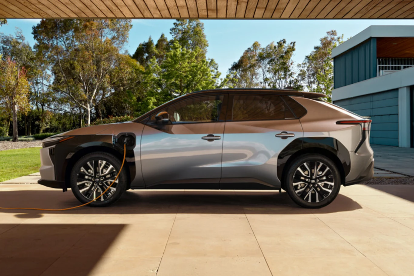Nowoczesny samochód elektryczny SUV w brązowo-szarym kolorze stoi pod zadaszeniem i jest podłączony do ładowarki. W tle widoczny ogród, wysokie drzewa oraz nowoczesny budynek mieszkalny.