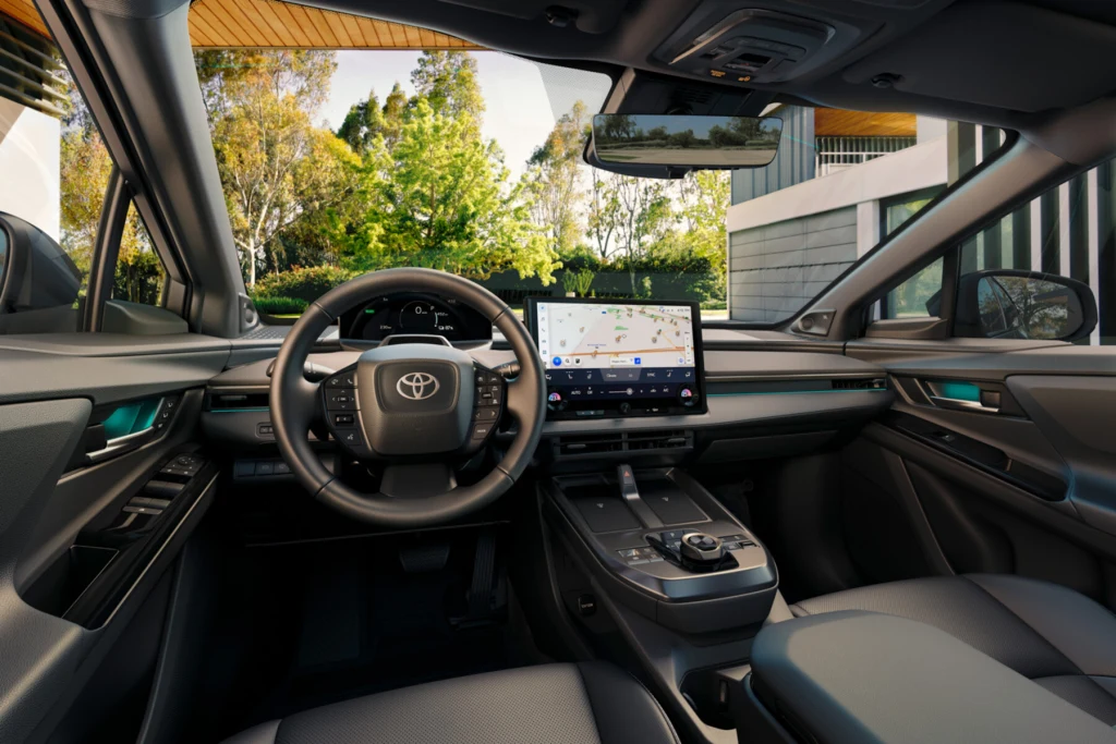 Wnętrze nowoczesnego samochodu marki Toyota z dużym wyświetlaczem centralnym, cyfrowym panelem kierowcy oraz elegancką deską rozdzielczą, siedzenia z wysokiej jakości materiałów, za oknami widoczna zieleń i nowoczesne budynki.