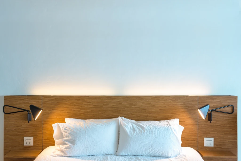 Nowoczesne, minimalistyczne łóżko z białą pościelą, podwójny zagłówek z drewna, po bokach dwie czarne lampki na ścianie, jasna i spokojna kolorystyka wnętrza.