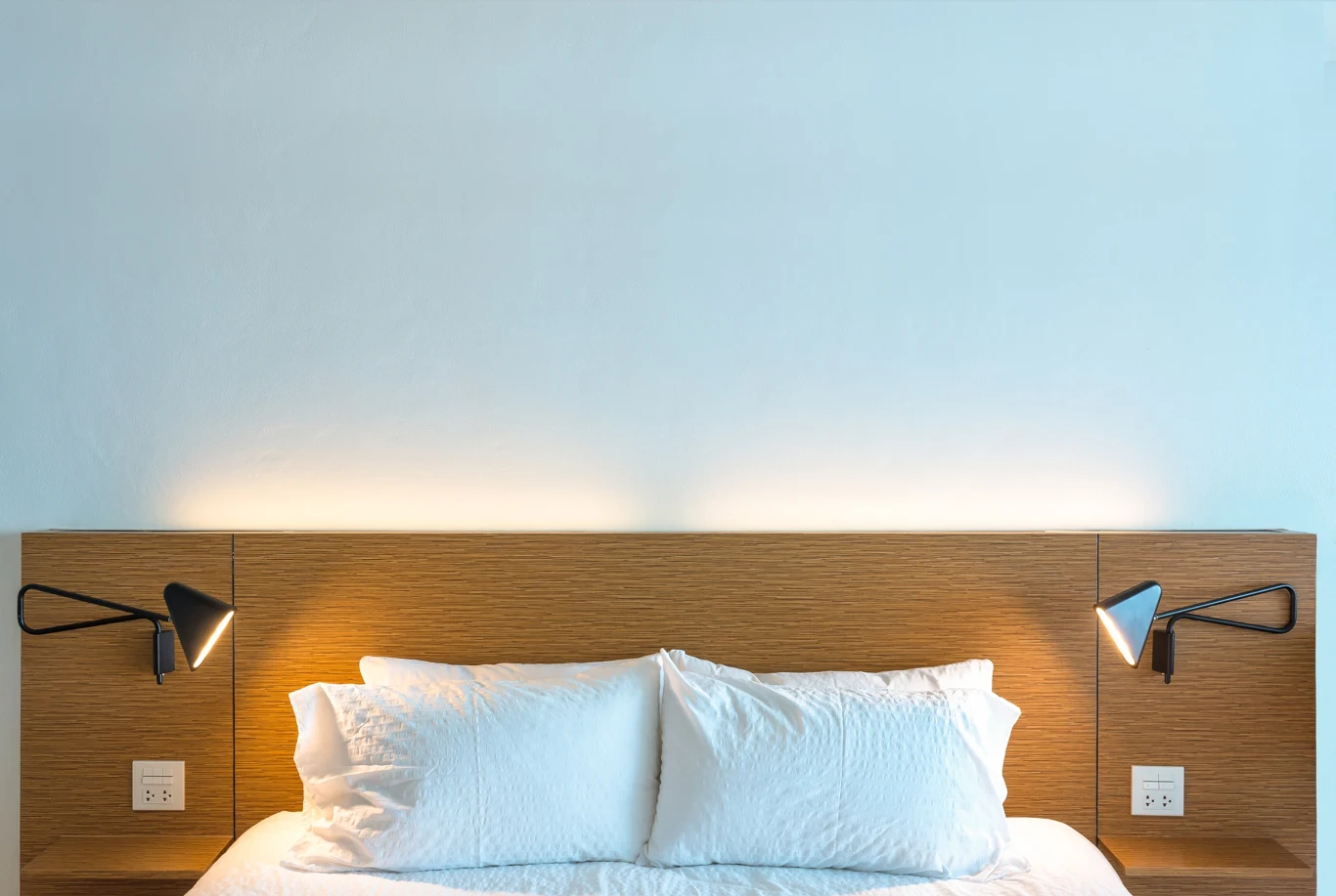 Nowoczesne, minimalistyczne łóżko z białą pościelą, podwójny zagłówek z drewna, po bokach dwie czarne lampki na ścianie, jasna i spokojna kolorystyka wnętrza.