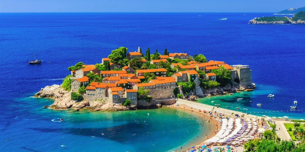 Wakacje w Czarnogorze w lato 2026 Malownicza wyspa z zabudowaniami o czerwonych dachach otoczona turkusowym morzem, u jej wybrzeży plaża z licznymi parasolami i turystami. W tle rozległa panorama lazurowej wody i odległe fragmenty lądu.
