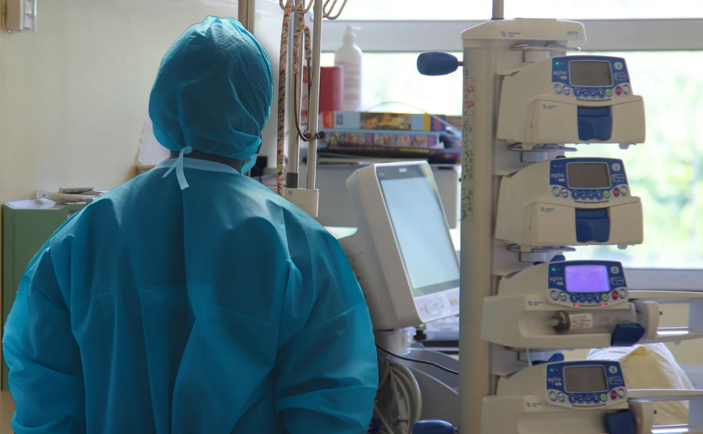 Osoba ubrana w ochronny kombinezon medyczny stoi w sali szpitalnej, otoczona zaawansowanym sprzętem medycznym i pompami infuzyjnymi, w tle widoczne okno dające naturalne światło.