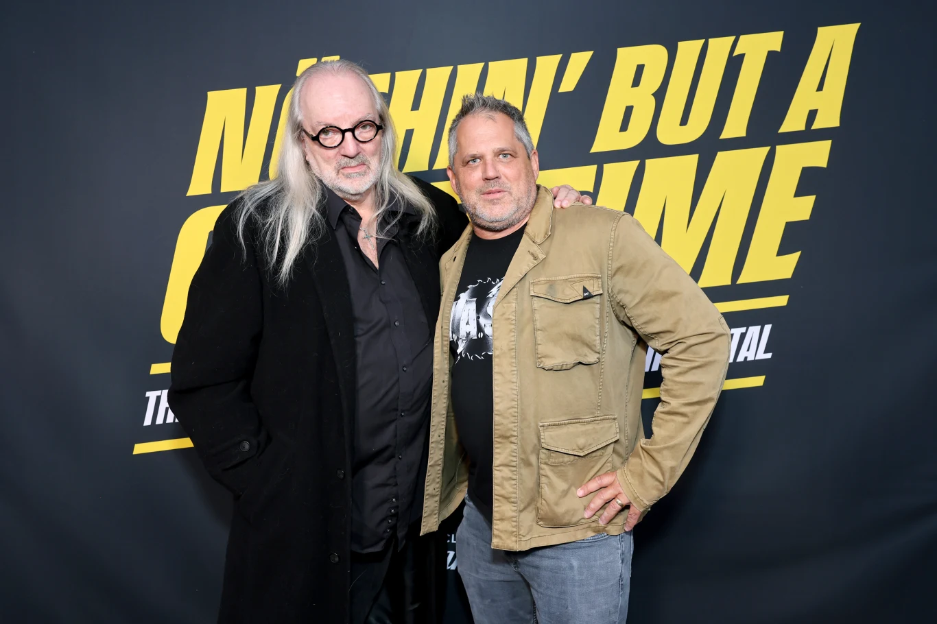 Dwóch mężczyzn pozuje wspólnie na tle czarnej ścianki z wyrazistym żółtym napisem, jeden w czarnym płaszczu i okrągłych okularach, drugi w jasnej kurtce z ręką opartą na ramieniu pierwszego mężczyzny.