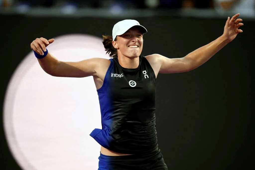 Iga Świątek wygrała WTA Finals 2023 Sportowiec ubrany w czarny strój sportowy i białą czapkę świętuje zwycięstwo na korcie tenisowym, unosząc ręce i uśmiechając się triumfalnie.