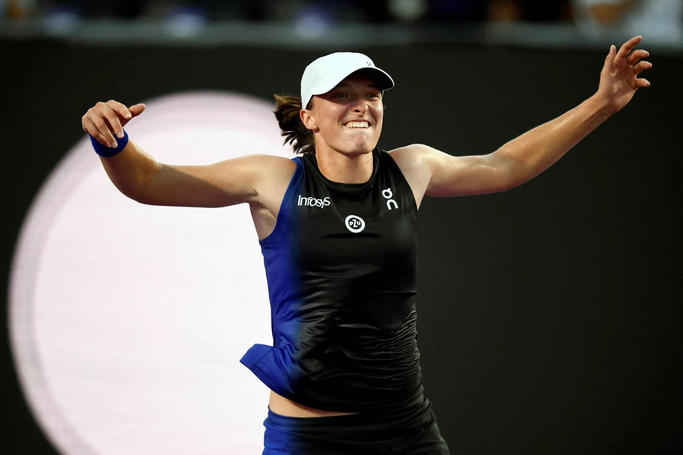 Iga Świątek wygrała WTA Finals 2023 Sportowiec ubrany w czarny strój sportowy i białą czapkę świętuje zwycięstwo na korcie tenisowym, unosząc ręce i uśmiechając się triumfalnie.