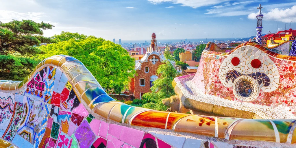 Barwne mozaikowe ławki i fantazyjna architektura otoczone bujną zielenią z panoramą miasta w tle, charakterystyczny styl katalońskiego modernizmu.