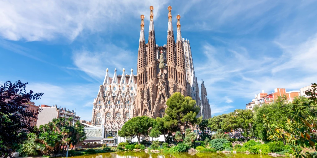 Bazylika Sagrada Familia w Barcelonie otoczona zielenią i błękitnym niebem, widoczna fasada z charakterystycznymi wieżami i detalami architektonicznymi.