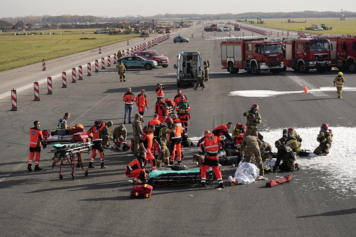 Ćwiczenia służb ratowniczych w Porcie Lotniczym Wrocław Grupa ratowników medycznych, strażaków i wojskowych udziela pomocy rannym na pasie startowym lotniska, wokół nich rozmieszczone nosze oraz sprzęt ratunkowy, w tle widoczne wozy strażackie i służbowe pojazdy.