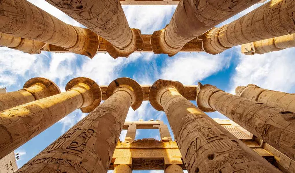Wysokie kamienne kolumny ozdobione hieroglifami widziane od dołu na tle nieba z delikatnymi chmurami, tworzące monumentalną strukturę starożytnej architektury.