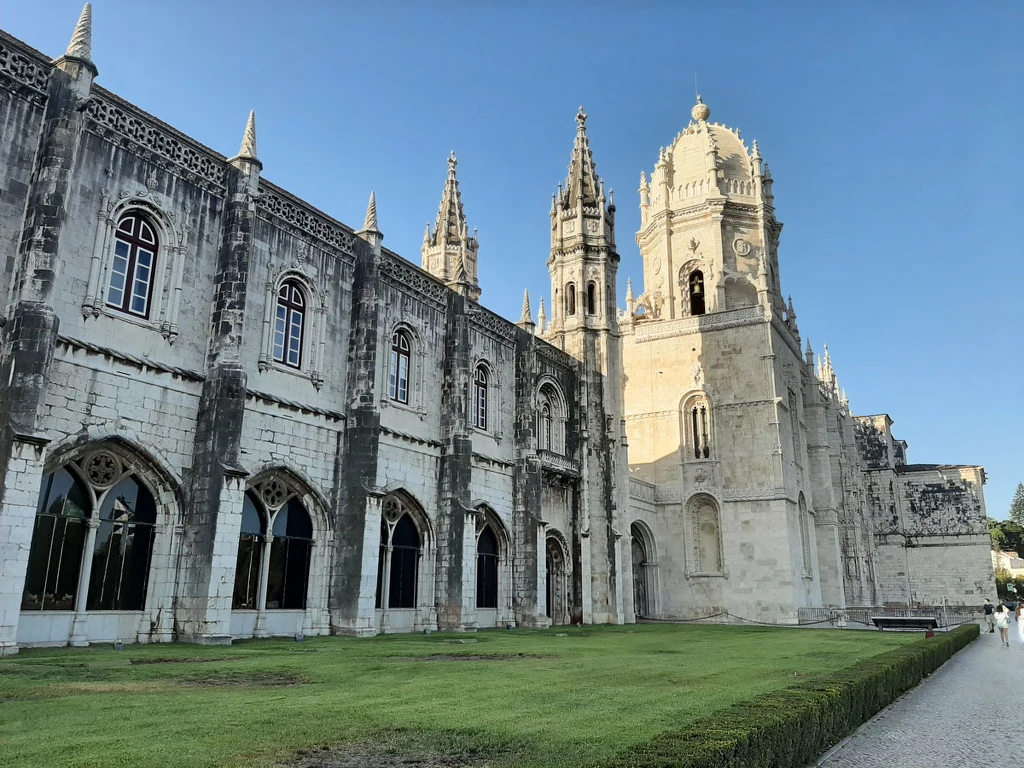 Gotycka fasada klasztoru z licznymi witrażowymi oknami, ozdobnymi łukami oraz wysokimi wieżami. Jasny kamień budynku kontrastuje z zielonym trawnikiem i błękitnym niebem, podkreślając majestat konstrukcji.