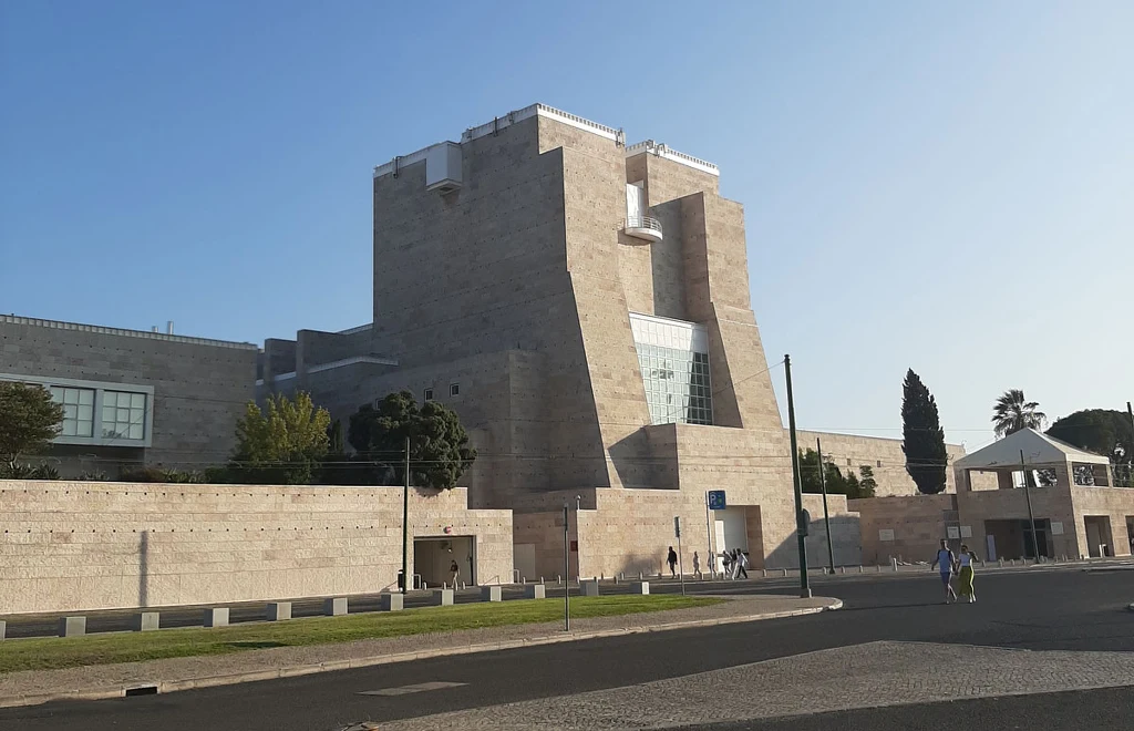 Nowoczesny, masywny budynek z jasnego kamienia, otoczony zielenią, z dużymi przeszkleniami i prostą architekturą. W tle widoczny jest kolejny jasny budynek oraz kilka osób poruszających się po placu przed budynkiem.