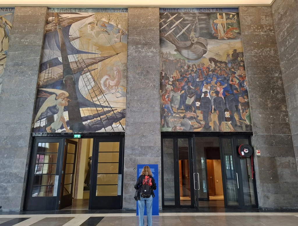 Kobieta z plecakiem stoi naprzeciwko dwóch wielkoformatowych malowideł ściennych umieszczonych ponad wejściami w monumentalnym wnętrzu budynku. Malowidła przedstawiają sceny z ludźmi oraz elementami nawiązującymi do postępu naukowego i eksploracji, z w...