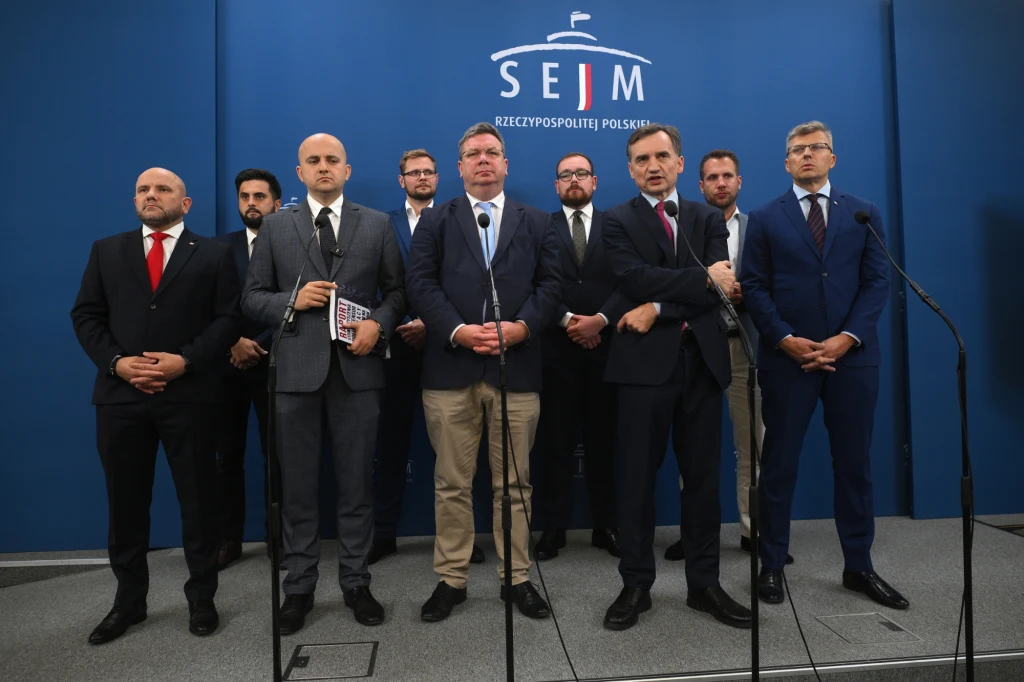 Grupa mężczyzn ubranych w oficjalne garnitury stoi na tle niebieskiej ściany z napisem SEJM Rzeczypospolitej Polskiej. Mężczyźni ustawieni są w kilku rzędach, większość patrzy prosto w obiektyw, część trzyma mikrofony, a jeden z nich trzyma notatnik.