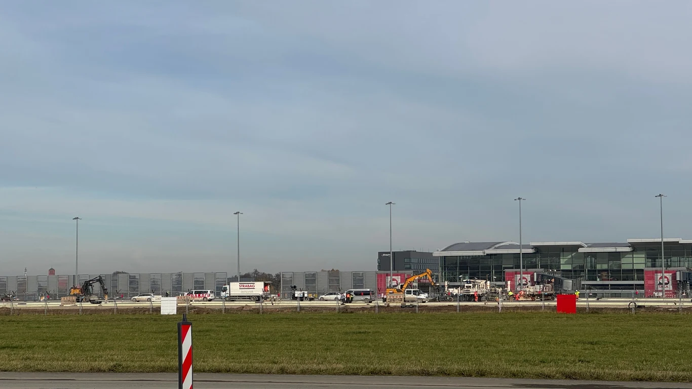 Trwa remont Portu Lotniczego Wrocław Plac budowy przy terminalu lotniska z widocznymi maszynami budowlanymi, samochodami i budynkiem terminala w tle, na pierwszym planie zielona trawa.