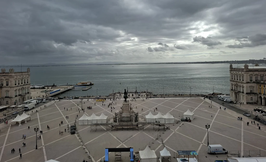 Rozległy plac miejski z marmurowym pomnikiem pośrodku, otoczony zabytkowymi budynkami. W tle widoczna zatoka, na niebie gęste chmury. Przestrzeń placu ozdabiają białe namioty, po placu spaceruje grupa ludzi.