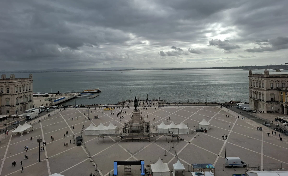 Rozległy plac miejski z marmurowym pomnikiem pośrodku, otoczony zabytkowymi budynkami. W tle widoczna zatoka, na niebie gęste chmury. Przestrzeń placu ozdabiają białe namioty, po placu spaceruje grupa ludzi.