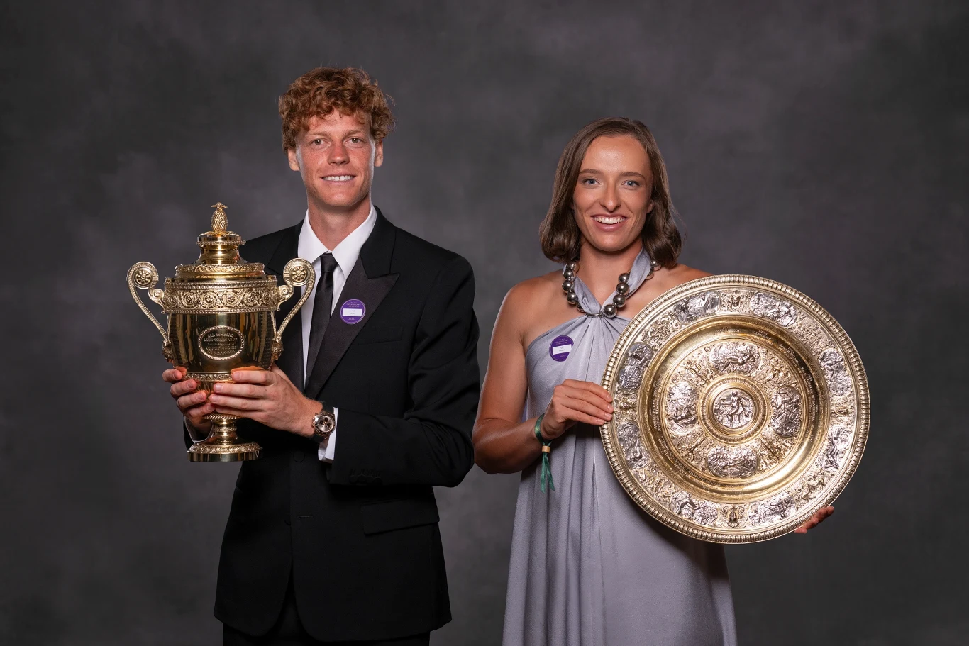 Jannik Sinner i Iga Świątek z trofeami za zwycięstwo w Wimbledonie Mężczyzna w garniturze oraz kobieta w eleganckiej sukni stoją razem na ciemnym tle, każdy z nich trzyma prestiżowe trofeum tenisowe – złoty puchar i srebrną paterę, oboje uśmiechnięci, w pozie oficjalnej.