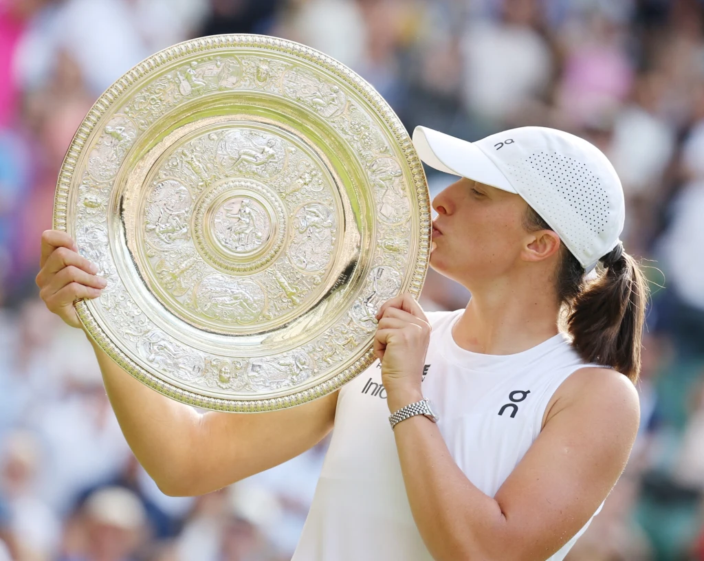 Iga Świątek z trofeum za wygraną w Wimbledonie Tenisistka ubrana na biało całuje duże srebrne trofeum, wyrażając radość i dumę ze zdobycia nagrody podczas ważnego turnieju tenisowego.