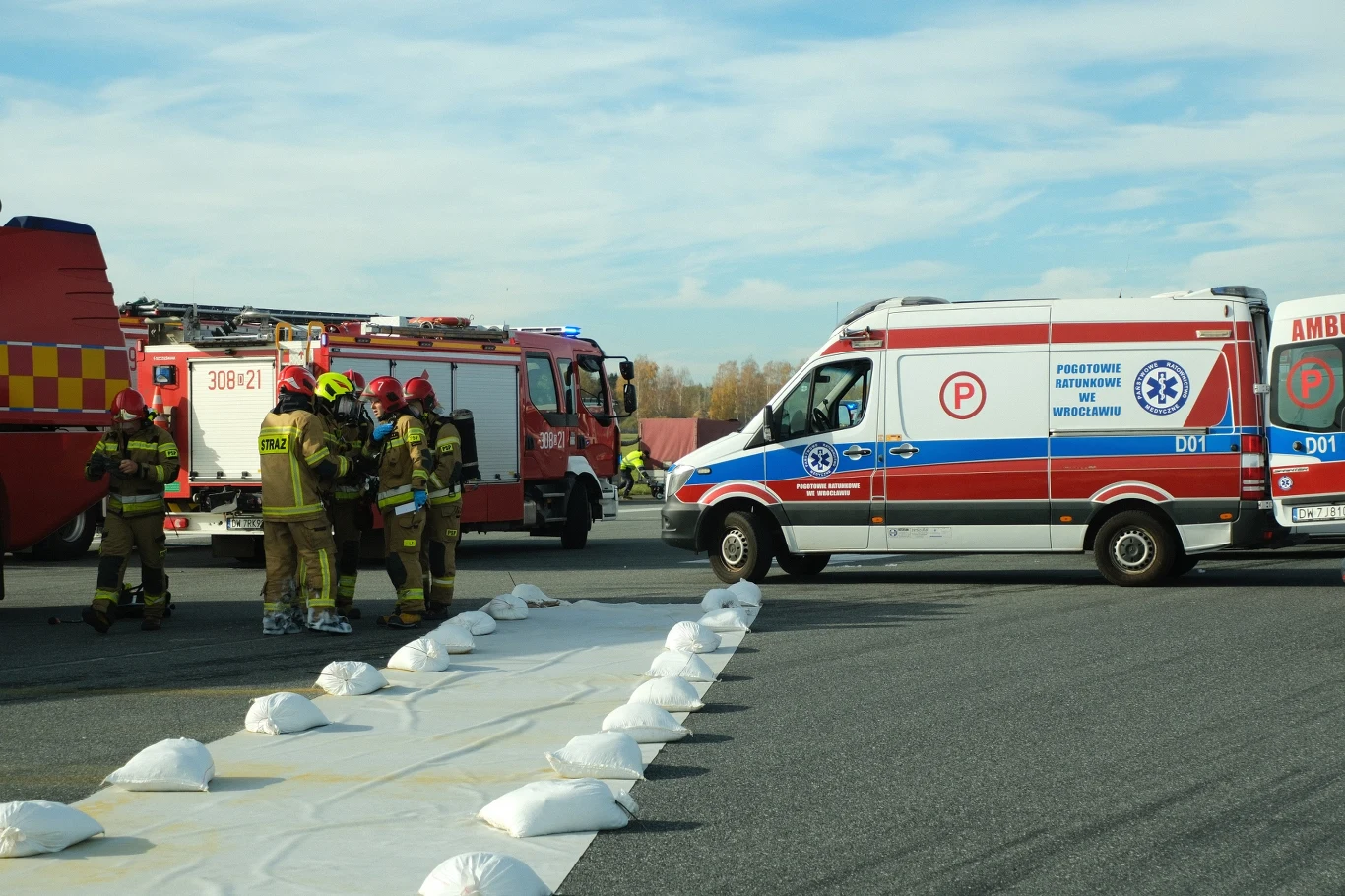 Liczne służby bardzo szybko stawiły się na miejscu zdarzenia. Strażacy w pełnym umundurowaniu stojący obok wozu strażackiego oraz karetki pogotowia, na asfalcie rozłożone białe worki prawdopodobnie zabezpieczające teren akcji ratowniczej, w tle widoczny fragment pojazdu strażackiego.