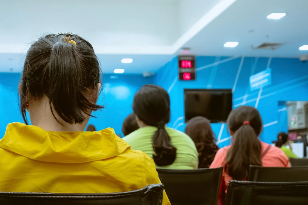 Osoby siedzące na krzesłach w poczekalni, widok z tyłu. Na ścianie znajduje się elektroniczny wyświetlacz z numerami, wnętrze jest jasne, utrzymane w niebiesko-białej kolorystyce.