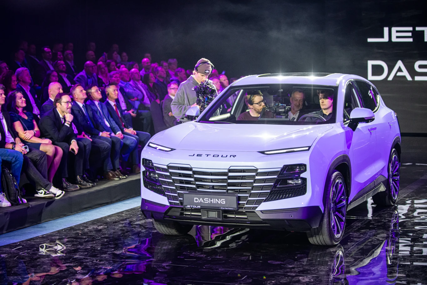 Nowoczesny samochód SUV marki Jetour Dashing stojący na scenie podczas oficjalnej prezentacji, otoczony przez widownię oraz kamerzystów, intensywne oświetlenie sceniczne podkreśla kształty pojazdu.