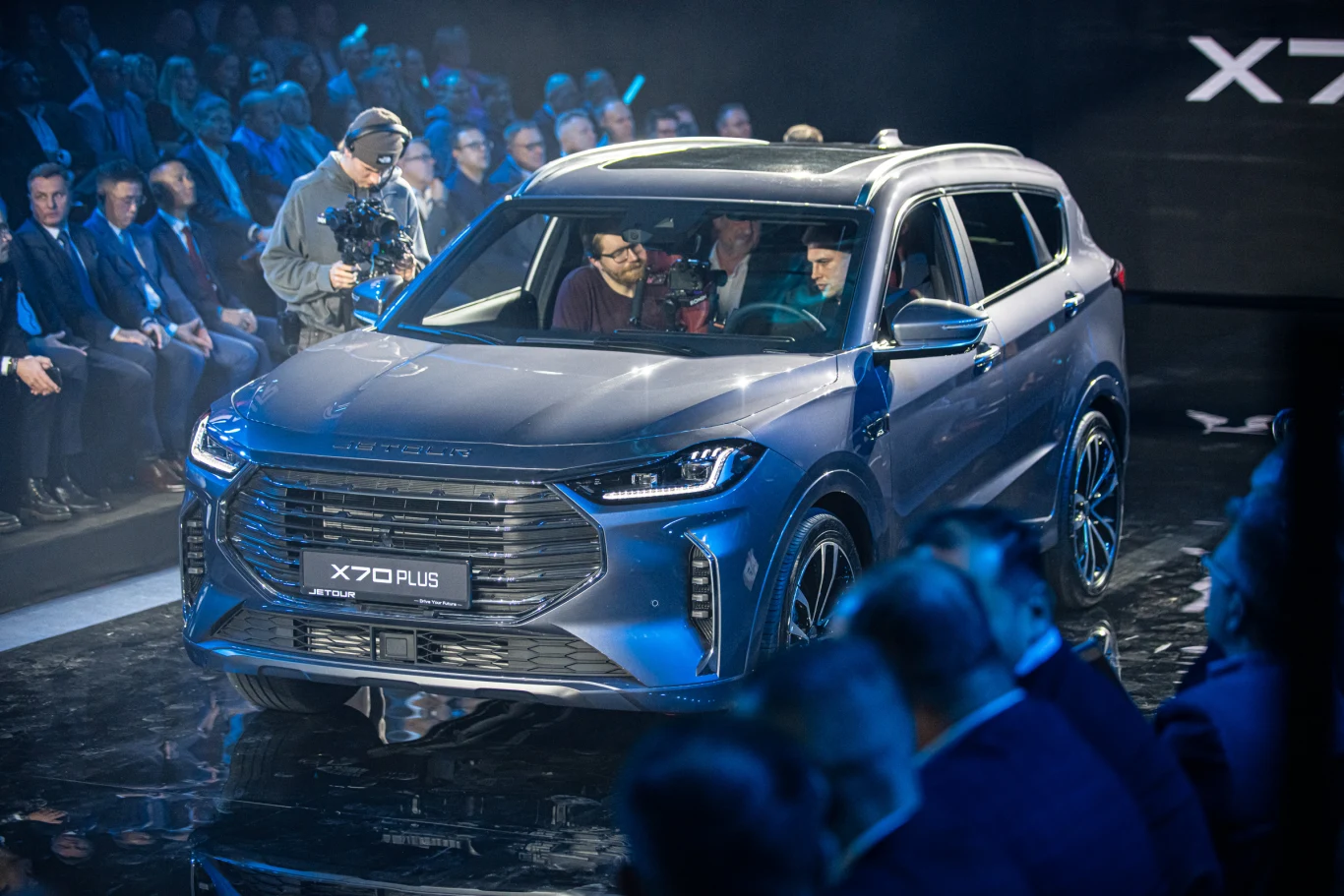 Nowoczesny srebrny SUV X70 Plus podczas oficjalnej premiery, otoczony przez widownię i dziennikarzy. Auto stoi na oświetlonej scenie, a z przodu, za kierownicą, siedzą dwie osoby. Tłum ludzi w garniturach śledzi wydarzenie, podkreślając rangę premiery.