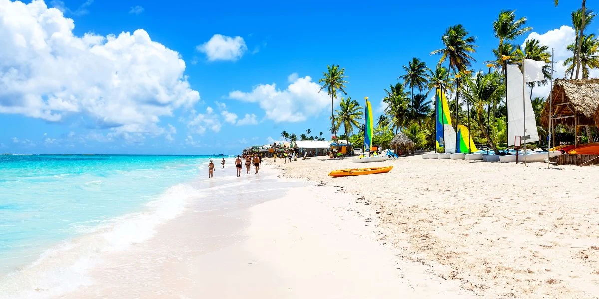 Szeroka, piaszczysta plaża z turkusową wodą, palmami oraz kilkoma osobami spacerującymi brzegiem. Po prawej stronie widoczne są żaglówki, sprzęt plażowy i niewielkie chatki, a całość oświetlona jest jasnym, słonecznym niebem.