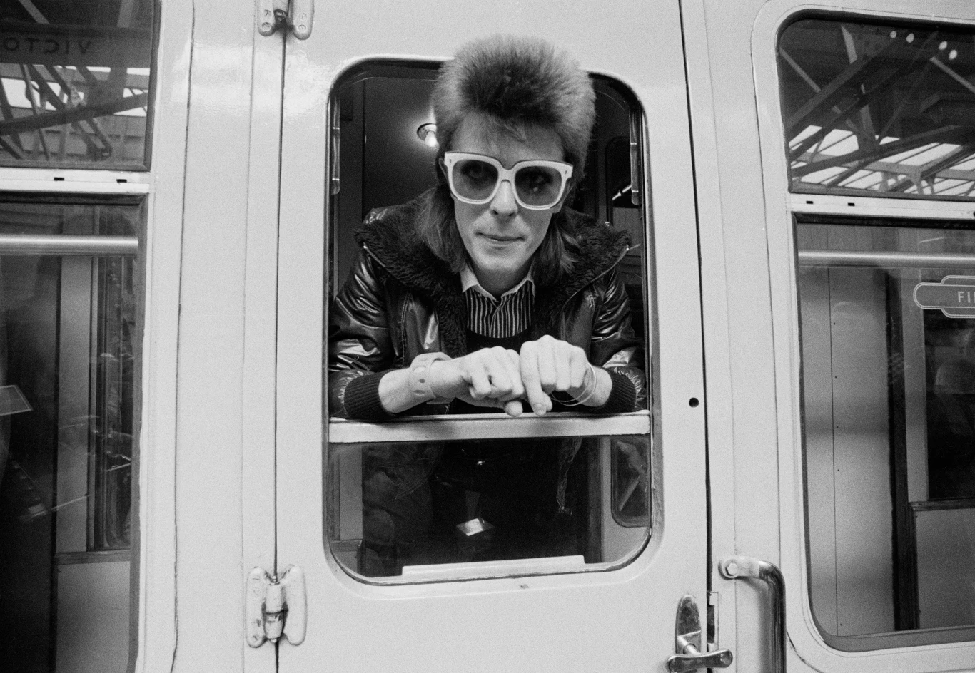 David Bowie w Londynie - 9 lipca 1973 r. Mężczyzna z charakterystyczną fryzurą i dużymi okularami przeciwsłonecznymi wychyla się przez otwarte okno wagonu pociągu, trzymając dłonie na ramie okna, patrząc bezpośrednio w stronę obiektywu.