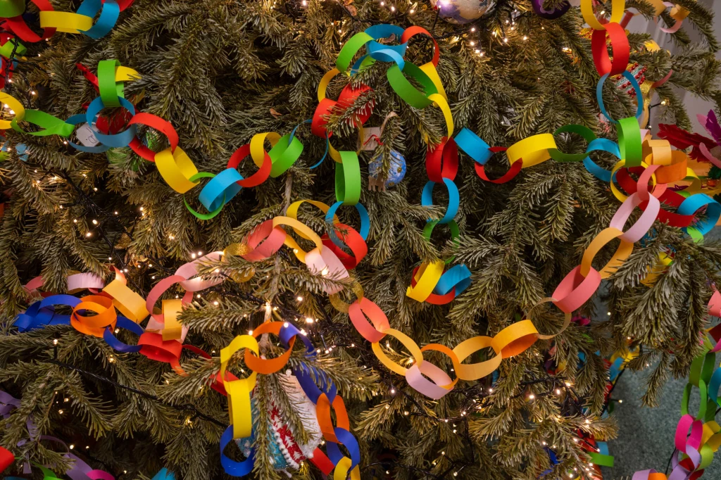 Ozdoby świąteczne DIY Kolorowy łańcuch z papierowych ogniw oplata gałęzie choinki, w tle widoczne drobne lampki świąteczne migoczące między igłami.