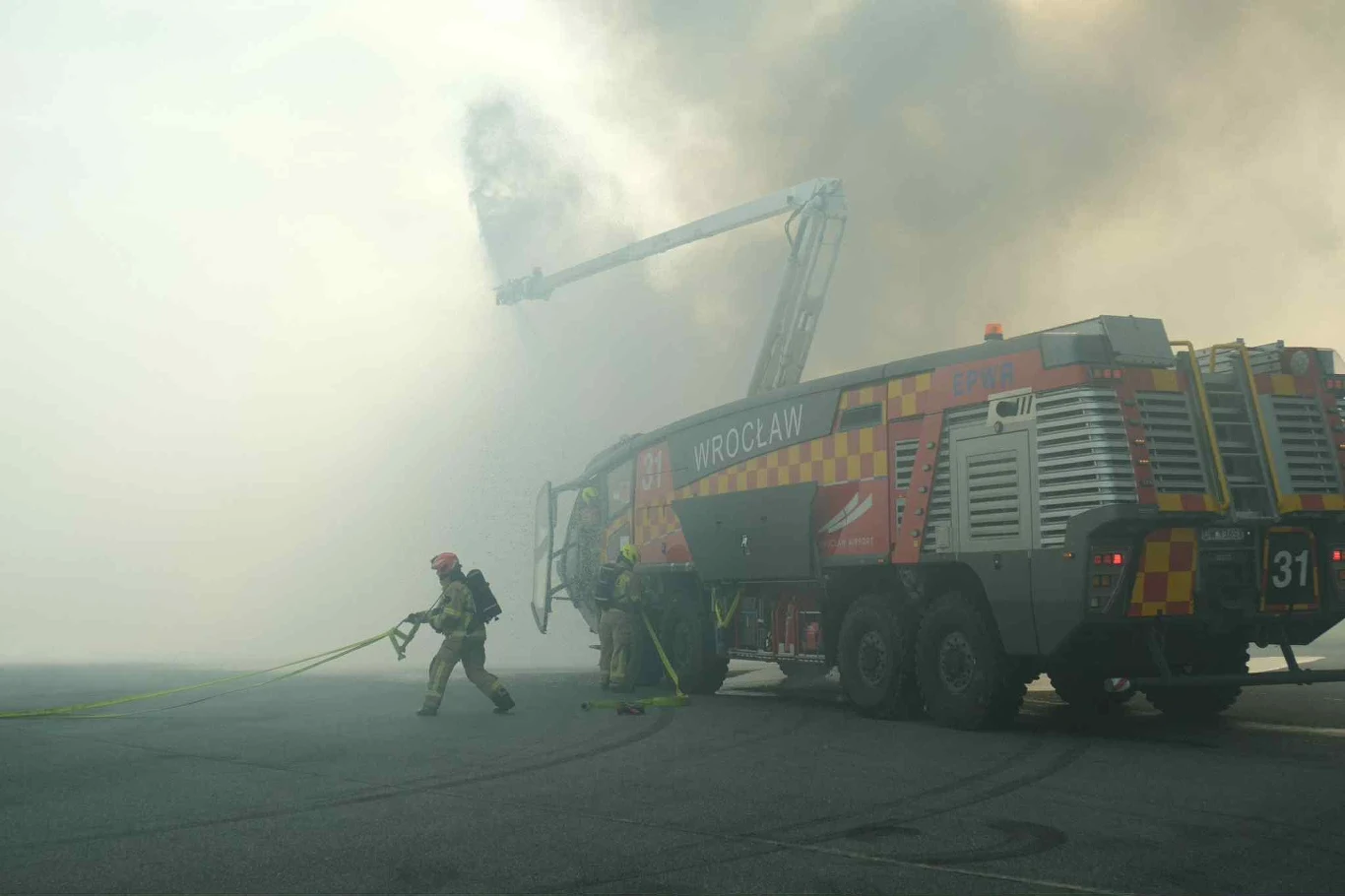 Widoczność była bardzo ograniczona - dla służb praca w takich warunkach to codzienność Strażacy w odzieży ochronnej obsługują wąż gaśniczy przy specjalistycznym pojeździe strażackim na tle gęstego dymu, z wysięgnikiem wodnym skierowanym w stronę zadymionego obszaru.