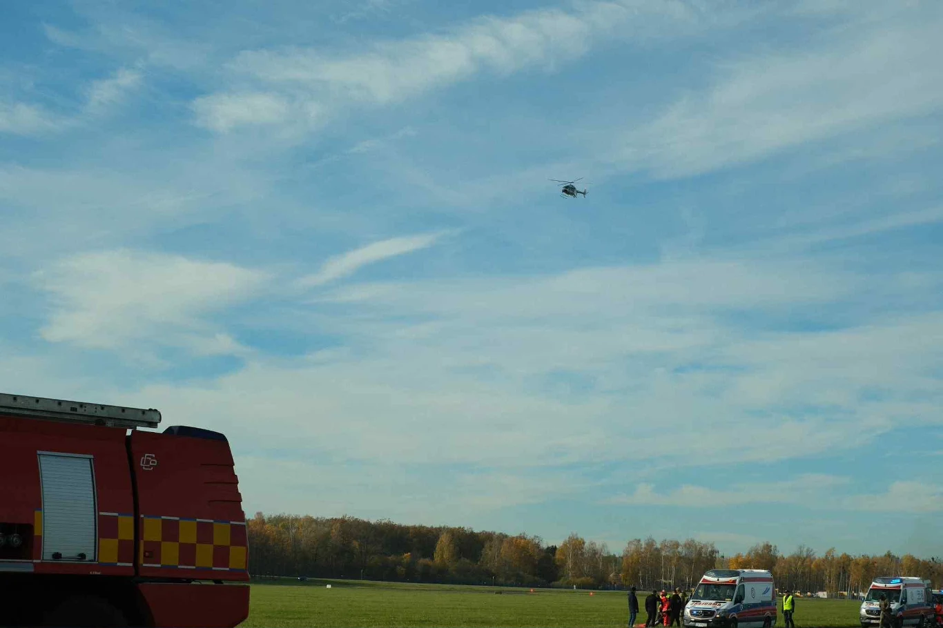 Nad naszymi głowami szybko pojawiły się śmigłowce Straż pożarna, ambulans, grupa osób w odzieży ochronnej oraz samochody ratownicze stoją na zielonym terenie, nad którym leci helikopter, a w tle rozciąga się las i niebo z lekkimi chmurami.