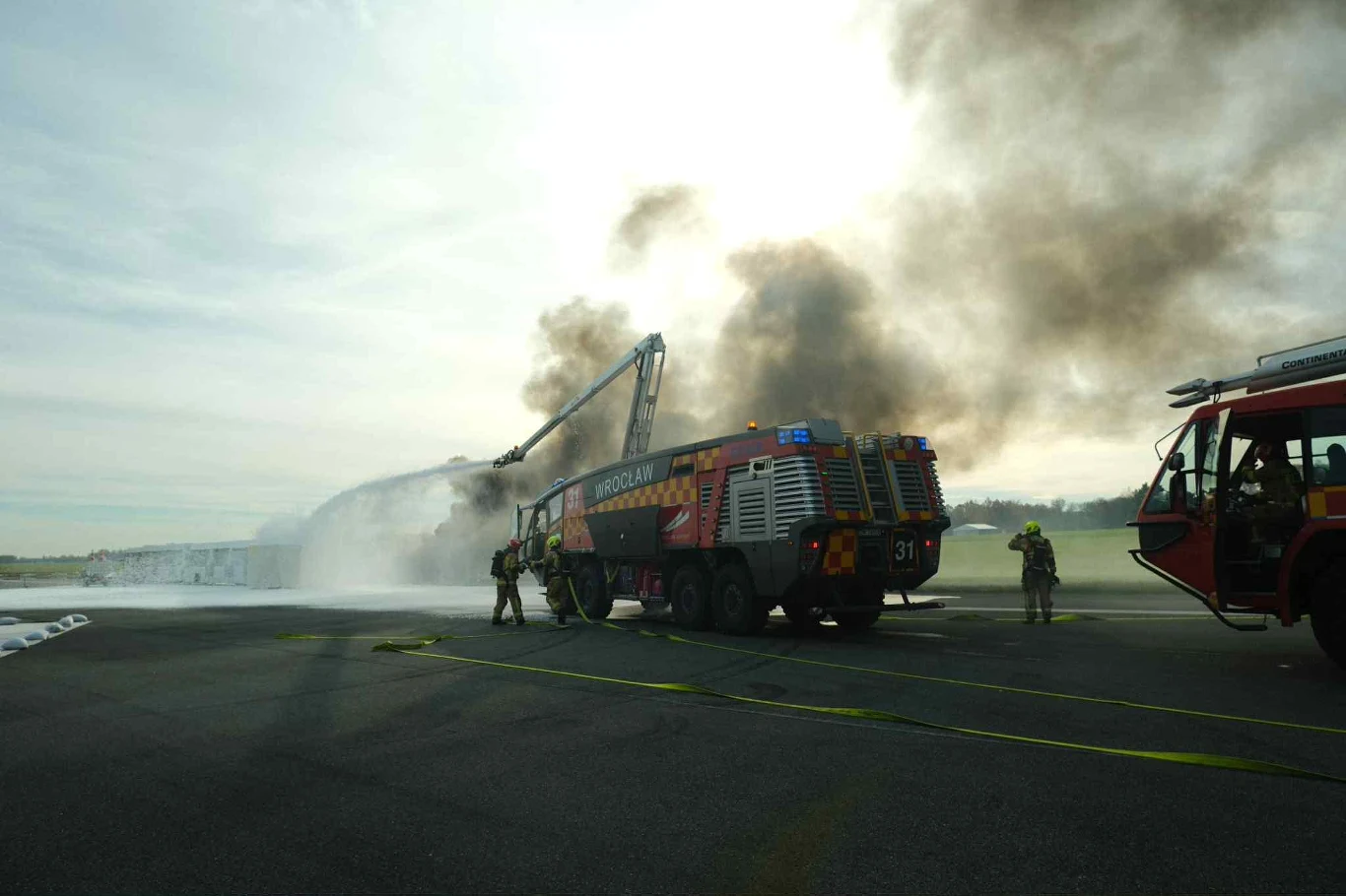 Dzięki sprawnemu działaniu służb, sytuację szybko dało się opanować Strażacy gaszący pożar przy użyciu specjalistycznych wozów strażackich, unoszący się gęsty dym na tle jasnego nieba, widoczne wyposażenie i intensywna akcja gaśnicza.