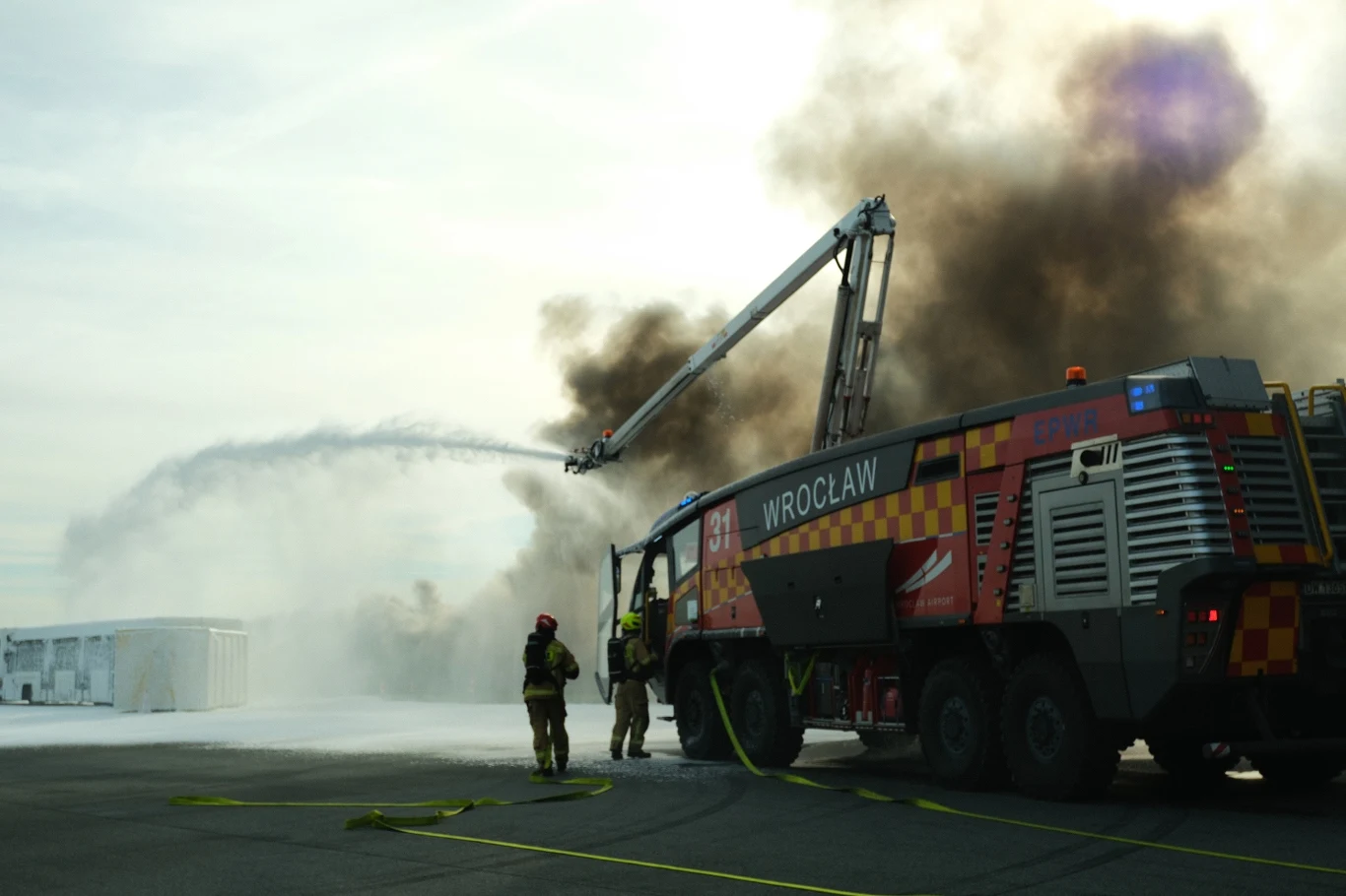 Ćwiczenia ratowniczo-gaśnicze w Porcie Lotniczym we Wrocławiu. Strażacki pojazd ratowniczy z napisem Wrocław gasi pożar gęstą pianą przy użyciu wysięgnika, wokół dym i kilku strażaków w kombinezonach ochronnych.