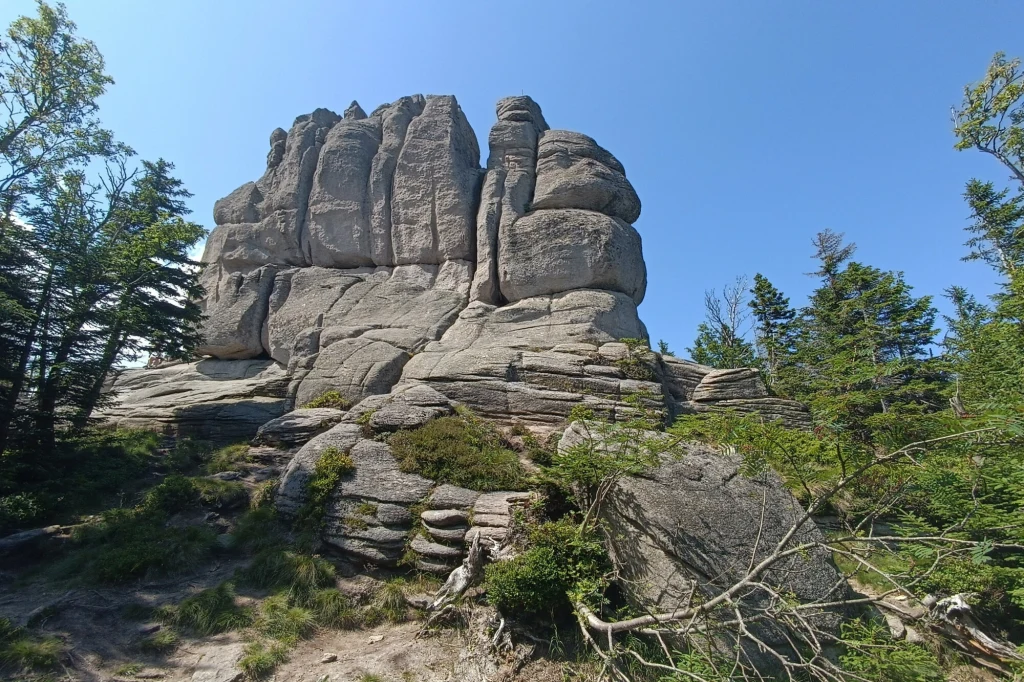 Wybierasz się na długi weekend w Karkonosze? Lepiej zapoznaj się z komunikatami KPN Monumentalna, naturalna formacja skalna otoczona zielonymi drzewami pod błękitnym niebem, z wyraźnymi warstwami i zaokrąglonymi kształtami skał.