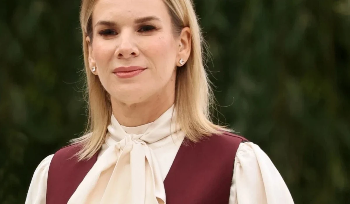 Marta Nawrocka Kobieta o jasnych włosach ubrana w elegancką białą bluzkę z wiązaniem pod szyją oraz bordową kamizelkę, tło ciemnozielone, rozmyte.