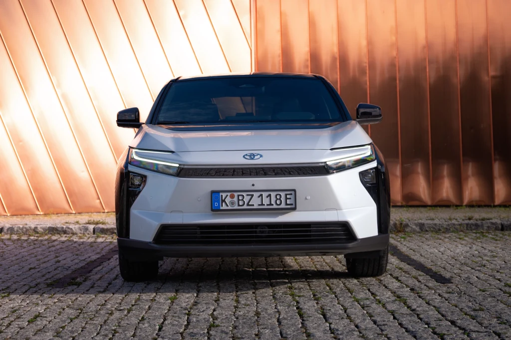 Nowoczesny, srebrny samochód osobowy marki Toyota ustawiony na brukowanym podłożu, na tle miedzianej, geometrycznej ściany. Auto na niemieckich tablicach rejestracyjnych, widok od frontu.