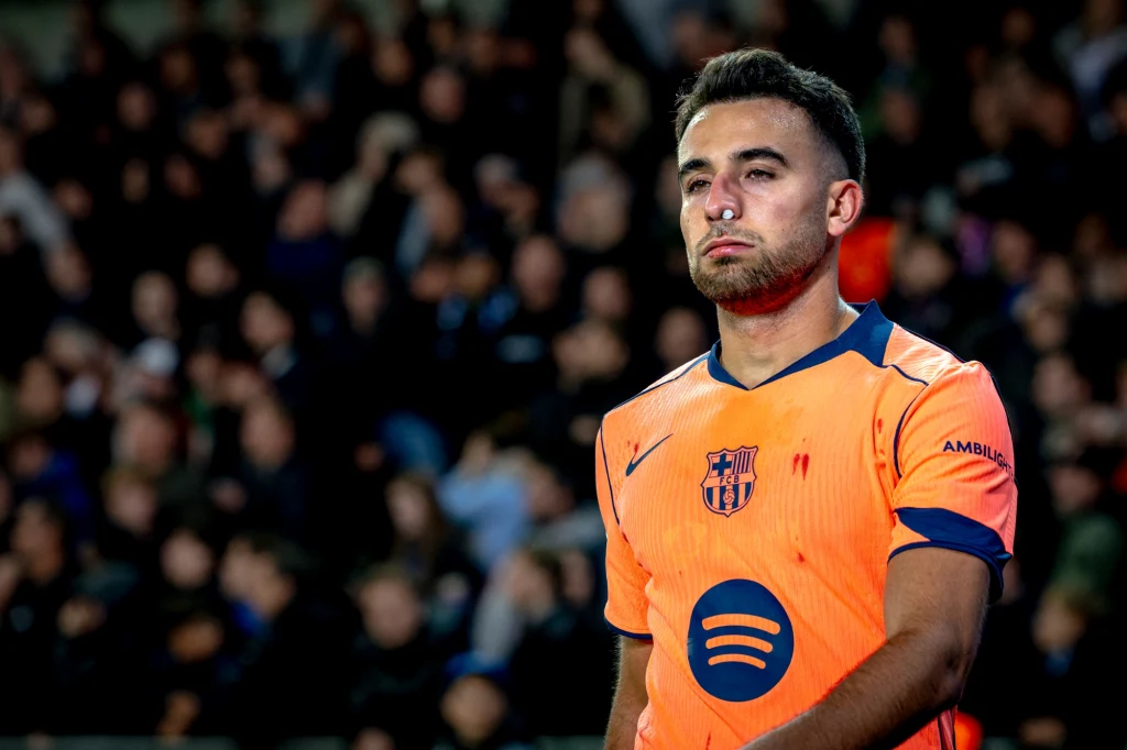 Młody piłkarz w pomarańczowej koszulce z herbem FC Barcelony i logo sponsora Spotify stoi na tle rozmytej publiczności, sprawiając wrażenie skupionego oraz odrobinę zmęczonego.