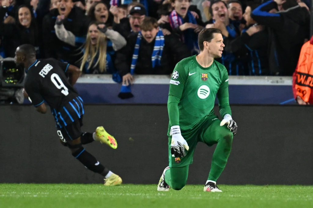 Piłkarz w zielonym stroju bramkarza klęczy na murawie, w tle zawodnik drużyny przeciwnej biegnie w kierunku kibiców, którzy reagują emocjonalnie swoim dopingiem