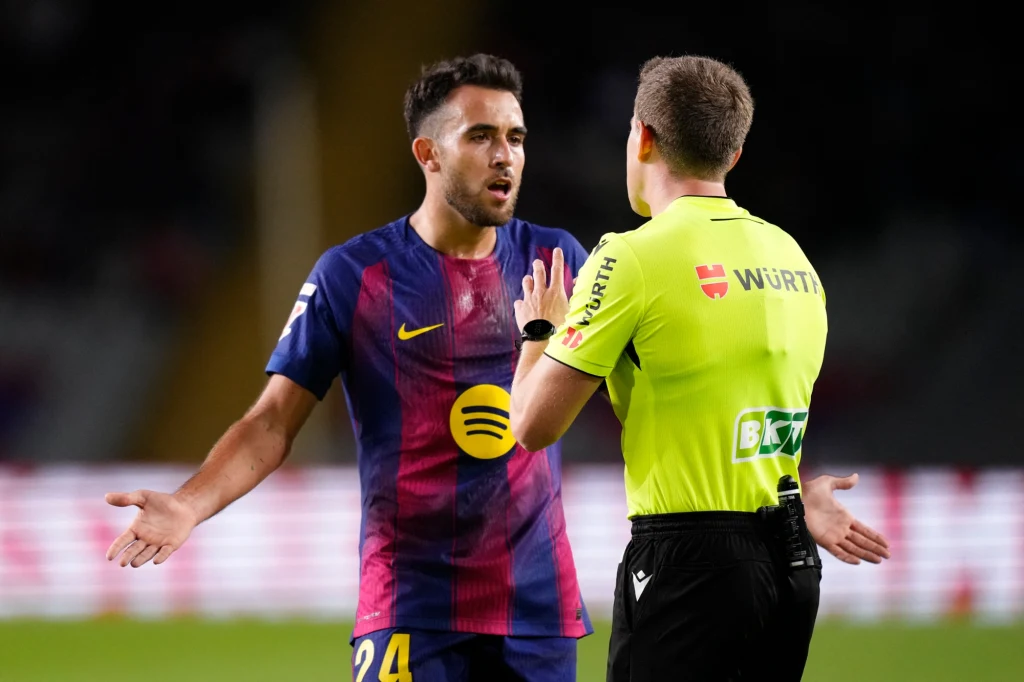 Piłkarz ubrany w strój drużyny FC Barcelona gestykuluje, dyskutując z sędzią w żółtej koszulce i czarnych spodenkach na boisku piłkarskim, wyrażając niezadowolenie lub zaskoczenie decyzją sędziego.