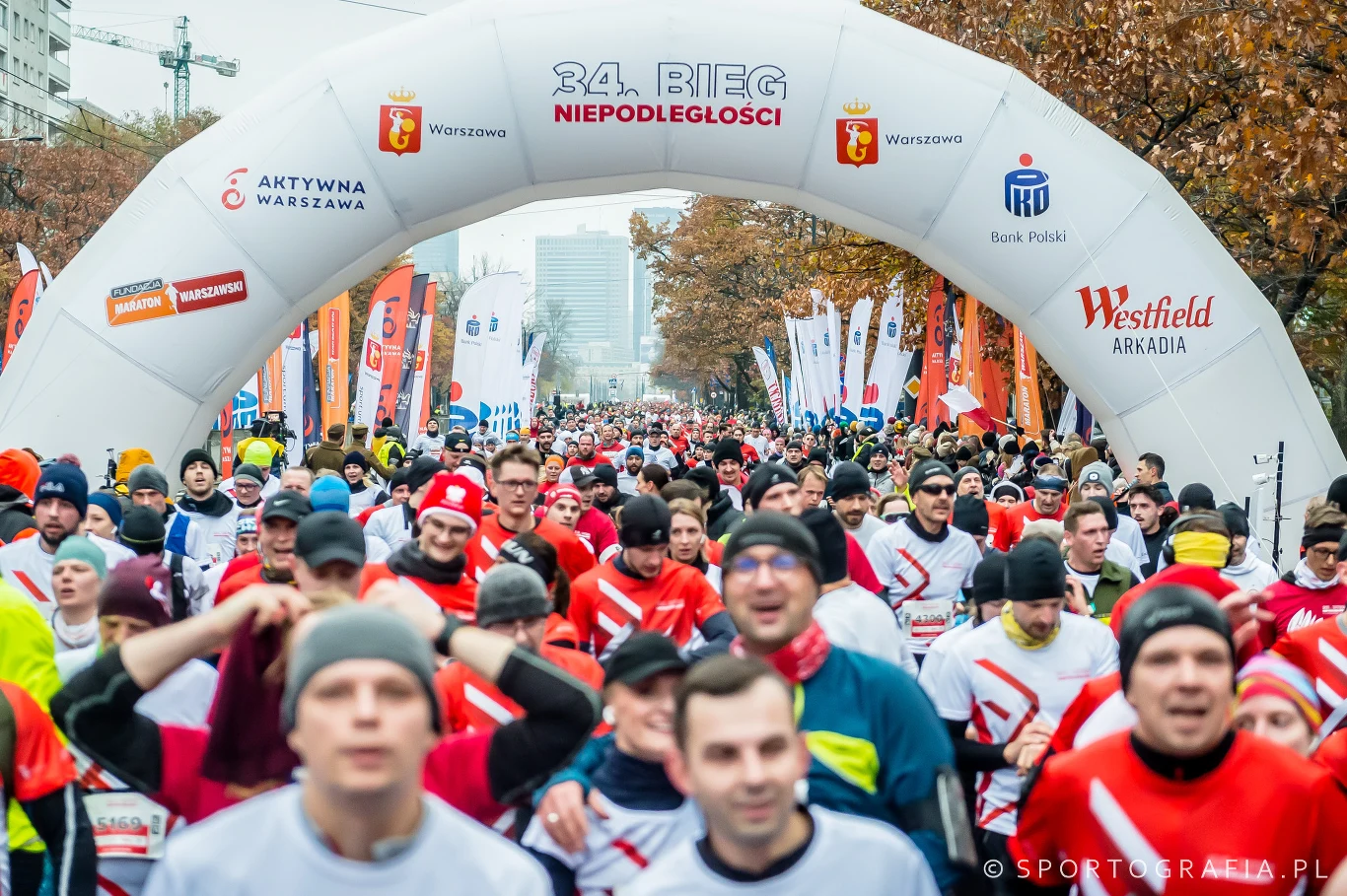 Tłum uczestników biegu ulicznego pod dużym łukiem startowym z napisem 34. Bieg Niepodległości, liczne flagi oraz bannery w tle, jesienna sceneria miejskiej alei.