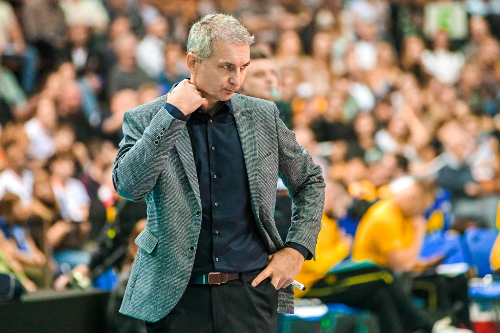 Mężczyzna w szarej marynarce i ciemnej koszuli stoi na tle tłumu kibiców, wykonuje gest głową i ręką, prawdopodobnie rozważając decyzję podczas wydarzenia sportowego, na trybunach widoczne osoby ubrane na żółto.