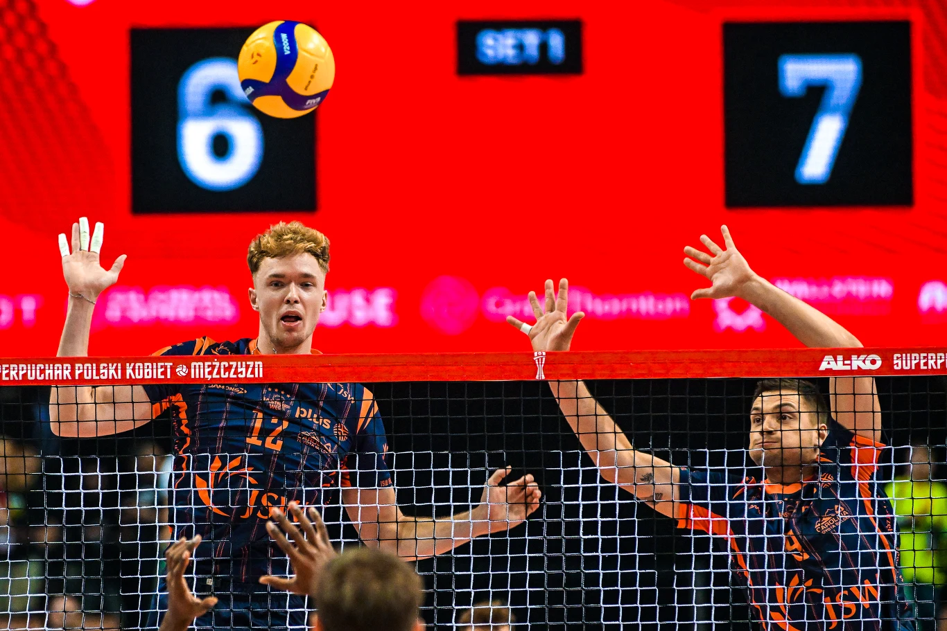 Dwóch siatkarzy ubranych w granatowo-pomarańczowe stroje wykonuje blok przy siatce podczas meczu, w tle widoczne są cyfry wyniku 6 i 7 oraz napis informujący o Superpucharze Polski.