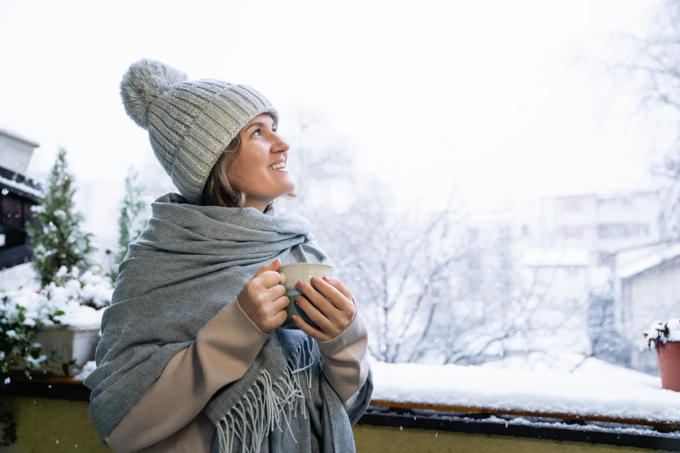W czym tkwi fiński sekret szczęścia? Młoda kobieta w ciepłej czapce i szalu trzyma filiżankę, stojąc na balkonie pokrytym śniegiem. W tle widoczne zabudowania i drzewa przysypane świeżym śniegiem.