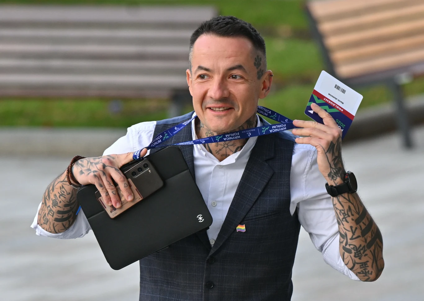 Uśmiechnięty mężczyzna z tatuażami na rękach trzyma w jednej dłoni identyfikator na niebieskiej smyczy, a w drugiej czarny tablet oraz telefon. Ubrany w białą koszulę i kamizelkę z przypinką w barwach Ukrainy, stoi na zewnątrz na tle ławek i zieleni.