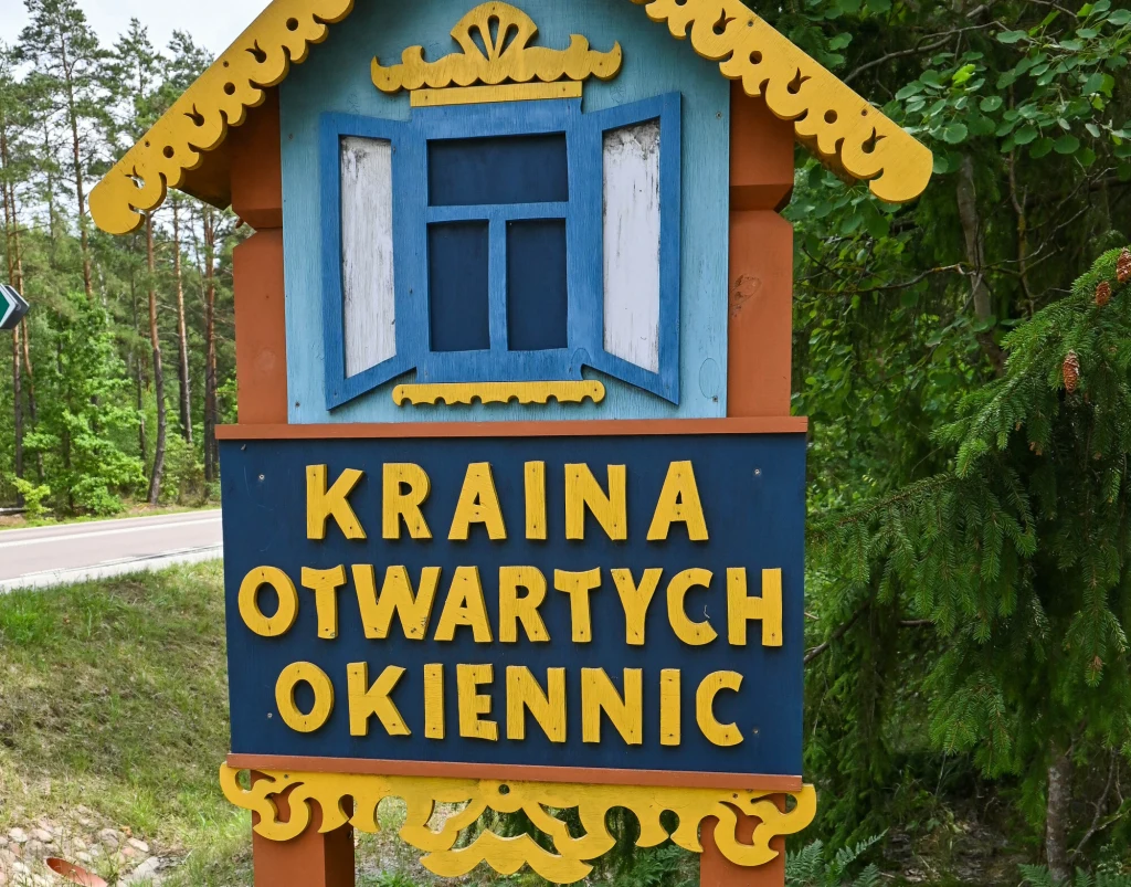 Kraina Otwartych Okiennic zaprasza na podróż w czasie Drewniany, kolorowy szyld z napisem Kraina Otwartych Okiennic, ozdobiony stylizowanym oknem, ustawiony przy drodze, na tle lasu.