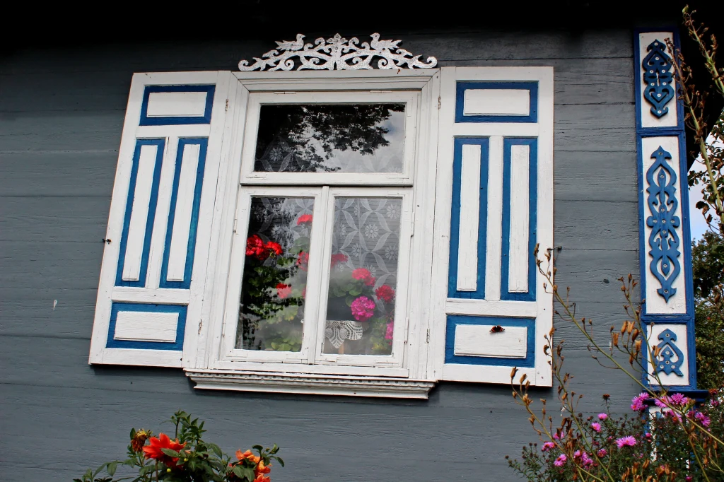 Każdy dom w Socach i okolicznych wioskach zachwyca unikatowymi zdobieniami Drewniane okno z dekoracyjnymi niebiesko-białymi okiennicami i koronkowymi firankami, przez szybę widoczne są czerwone i różowe pelargonie ustawione na parapecie, a wokół kwitną kolorowe kwiaty ogrodowe.