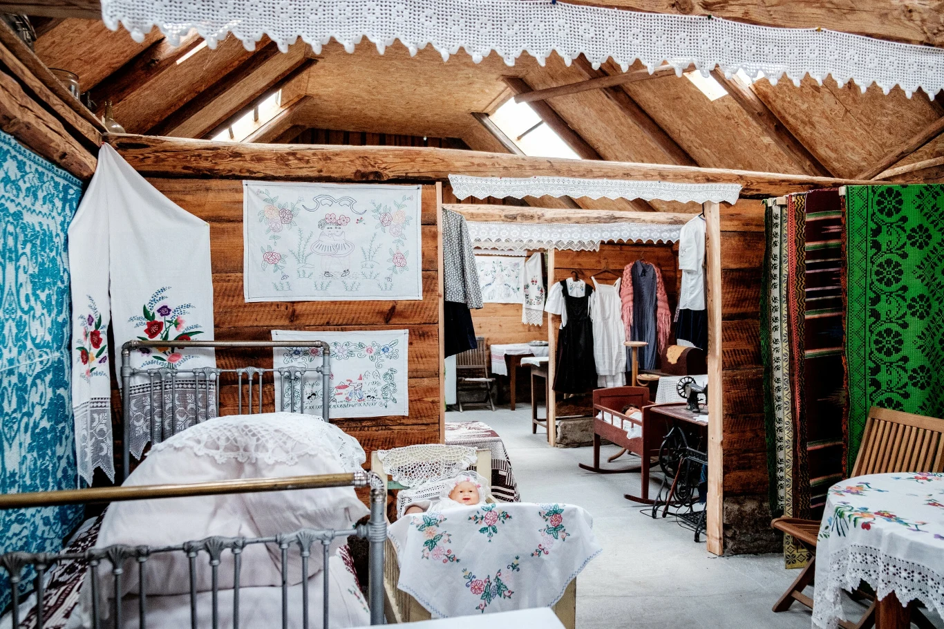 Tradycyjne wnętrze drewnianej izby z ręcznie haftowanymi obrusami, zasłonami i pościelą, dziecięcym łóżeczkiem, starodawnymi meblami oraz ludowymi strojami zawieszonymi na wieszakach.