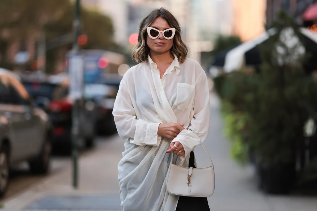 Sukienki maskujące brzuch Kobieta w stylowej białej koszuli i dużych okularach przeciwsłonecznych, trzymająca białą torebkę, spaceruje miejską ulicą.